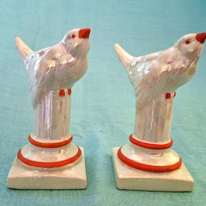May include: A pair of ceramic bird figurines featuring an iridescent white finish. Each bird is perched atop a cylindrical pedestal with a square base, accented by bright orange rings around the base and orange details on the beaks and feet.