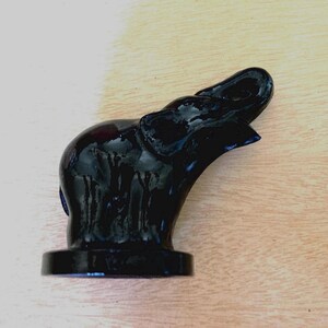 May include: A glossy black glass figurine of an elephant standing on a circular base. The elephant is depicted in profile with its trunk raised upward, featuring smooth, reflective surfaces and defined ear and leg contours in a solid black color.