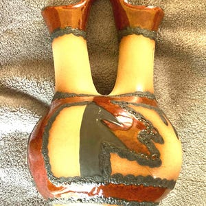 May include: A double-spouted ceramic vessel featuring a tan body with glossy reddish-brown accents on the rims and base. The piece is decorated with dark, textured, raised charcoal-colored lines that form geometric patterns across the surface.