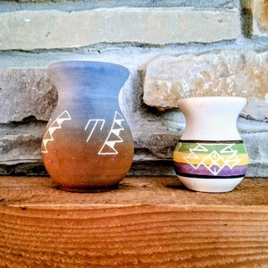 May include: Two small ceramic vases with flared rims. The larger vase features a blue-to-brown gradient finish with white geometric line patterns. The smaller white vase is decorated with horizontal green, yellow, and purple bands and white geometric designs.