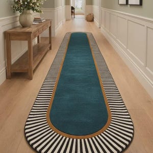 May include: A long, teal runner rug with a rounded end. The rug has a brown border and is framed by black and white striped edges. The rug is placed on a light wood floor in a hallway.