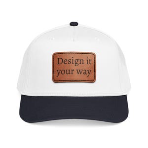 May include: A white baseball cap with a navy blue brim and a black button on top. The cap features a brown leather patch with the text "Design it your way".