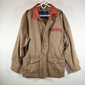 May include: A tan Structure brand jacket with a red collar and a red zippered chest pocket. The jacket has two front pockets with button closures and is hanging on a white hanger.