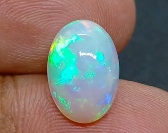 Könnte beinhalten: Ein ovaler Opal-Edelstein mit milchig-weißer Grundfarbe. Der Opal zeigt lebhafte Blitze in Grün, Blau und Gelb, wodurch ein Farbspiel-Effekt entsteht. Der Edelstein wird zwischen zwei Fingern gehalten.