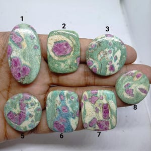 Peut inclure: Sept pierres précieuses cabochons polies présentant un mélange d'inclusions de rubis rose magenta vif incrustées dans une matrice de zoïsite vert pâle et crème. Chaque pierre est numérotée de 1 à 8, excluant le 4, avec des motifs uniques.