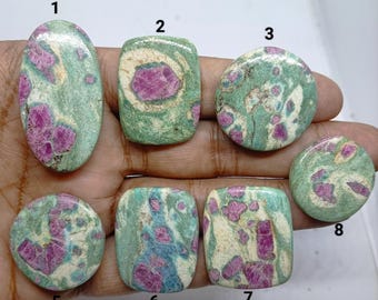 May include: Seven polished cabochon gemstones featuring a mix of vibrant magenta-pink ruby inclusions embedded within a pale green and cream-colored zoisite matrix. Each stone is numbered from 1 to 8, excluding 4, and displays unique patterns.