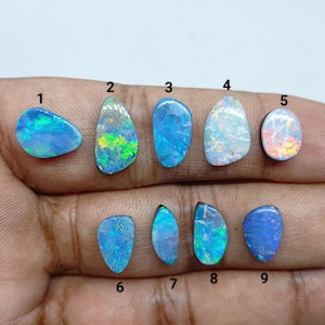 Pode incluir: Nove pedras preciosas de opala polidas dispostas em duas filas, cada uma rotulada com um número preto de um a nove. As pedras exibem flashes iridescentes de azul, verde, branco e laranja, com formas variadas e superfícies lisas.