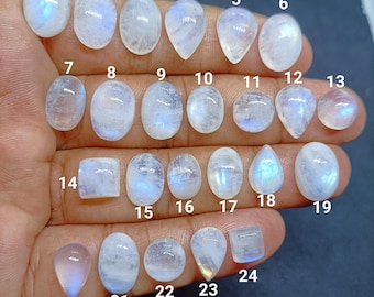May include: A collection of 24 polished moonstone gemstones in various shapes, including oval, pear, and square. The stones display a milky white base with blue and iridescent flashes. Each stone is numbered for identification.