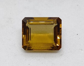 Op de afbeelding: Een rechthoekige edelsteen met een smaragdgeslepen vorm en een transparante, goudgele kleur. De steen heeft nauwkeurige, gefacetteerde randen die een geometrisch patroon van lichte en donkere reflecties op het oppervlak creëren.