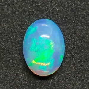 Puede incluir: Una piedra preciosa de ópalo de forma ovalada, con una base azul lechoso translúcido. La piedra presenta destellos vibrantes de verde y amarillo, creando un juego de colores. El ópalo se encuentra sobre un fondo oscuro y texturizado.
