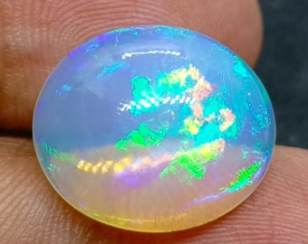 以下が含まれることがあります： 丸く虹色のオパール宝石。オパールは、緑、青、黄色のきらめきを持つ乳白色のベースカラーを示しています。宝石は滑らかで磨かれており、微妙な輝きで光を反射します。オパールは指の間に保持されています。