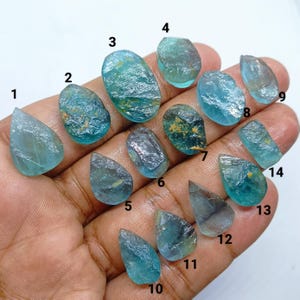 May include: A collection of fourteen raw, aquamarine-colored gemstones. The stones vary in shape, from teardrop to oval, and display a range of blue-green hues. Some stones have visible inclusions and textures, while others are more translucent.