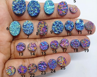 May include: A collection of iridescent druzy gemstones in various shapes and sizes. The gemstones display a range of colors, including blues, purples, and golds. The stones are oval and round, with a sparkling, crystalline texture.