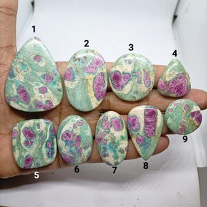 May include: Nine polished cabochon gemstones featuring a mix of vibrant magenta-pink inclusions embedded within a light green and white matrix. Each stone is numbered from one to nine, displaying unique organic patterns and smooth, rounded surfaces.