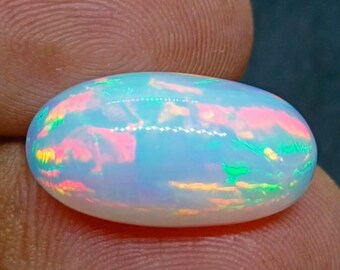 Könnte beinhalten: Ein ovaler Opal-Edelstein. Der Opal zeigt eine milchig-weiße Grundfarbe mit schillernden Blitzen in Rosa, Grün und Gelb. Der Edelstein wird zwischen zwei Fingern gehalten, wodurch seine polierte Oberfläche und das Farbspiel hervorgehoben werden.