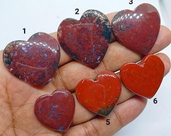 May include: Six heart-shaped gemstones in shades of red, some with green and white patterns. The polished stones are held in a hand, displaying their smooth surfaces and unique colour variations.