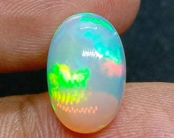 以下が含まれることがあります： 鮮やかな色の遊びを見せる、楕円形のオパール宝石。石は、半透明の乳白色のベースの中に、緑、青、ピンク、黄色の閃光を示しています。 オパールは指の間に保持され、その研磨された表面が示されています。