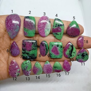 Può includere: Una collezione di diciotto gemme di rubino in zoisite lucidate di varie forme e dimensioni, ciascuna etichettata con un numero nero da 1 a 18. Le pietre mostrano un mix di vivaci inclusioni di rubino magenta e una matrice di zoisite verde.