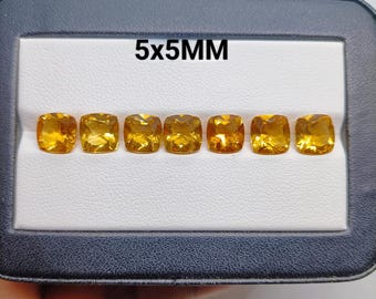 以下が含まれることがあります： 黒いディスプレイボックスに一列に並べられた、クッションカットの黄色いシトリンの宝石7個。テキストに示されている通り、各宝石のサイズは5x5mmです。石は透明で、光を反射するファセット加工された表面が、鮮やかな黄金色の輝きを放っています。