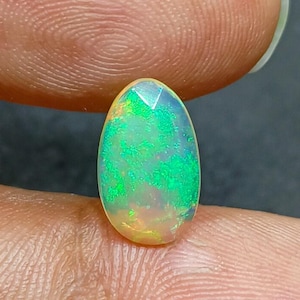 Peut inclure: Une pierre précieuse opale ovale. L'opale présente un mélange de couleurs vives, notamment du vert, du jaune et des touches d'orange. La surface polie de la pierre est mise en valeur par les doigts, révélant son jeu de couleurs interne.