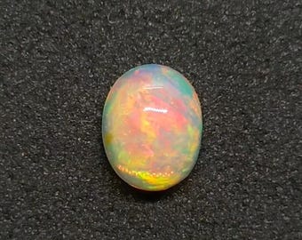 Könnte beinhalten: Ein ovaler Opal-Edelstein mit einer lebendigen Farbpalette. Der Opal zeigt eine Mischung aus gelben, orangen, rosa und grünen Farbtönen, die einen schimmernden Effekt erzeugen. Der Edelstein ist vor einem dunklen Hintergrund platziert.