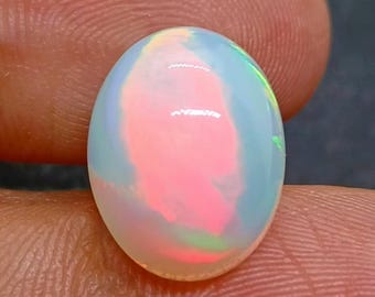 以下が含まれることがあります： 楕円形のオパール宝石。ピンク、青、緑の虹色の閃光で色の遊びを表現しています。研磨された表面は光を反射し、石の内部構造を示しています。宝石は指の間に挟まれています。