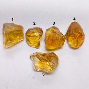 Puede incluir: Cinco cristales de citrino en bruto, translúcidos y de color amarillo dorado, dispuestos en grupo. Cada piedra presenta bordes irregulares y dentados con una textura cristalina facetada. Las piedras están numeradas con los dígitos negros 1, 2, 3, 4 y 5.
