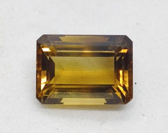 May include: A rectangular, emerald-cut gemstone with a deep golden-brown hue. The stone features faceted surfaces that create internal light reflections and shadows, showcasing a transparent, warm amber colour throughout its geometric structure.