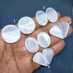 Puede incluir: Una colección de cabujones de selenita blanca pulida en varias formas, incluyendo círculos, óvalos y gotas. Las piedras están numeradas del 1 al 5, mostrando su textura suave, translúcida y fibrosa bajo la luz natural.