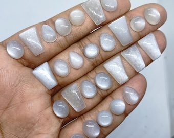 May include: A collection of polished, translucent white moonstone cabochons in various shapes, including round, teardrop, and trapezoidal forms, displayed across the fingers of a hand to showcase their smooth, reflective surfaces.