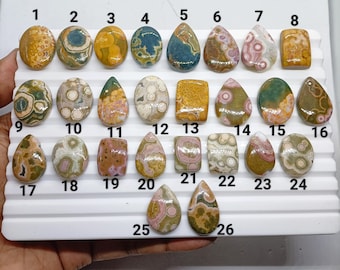 May include: A collection of twenty-six polished, multi-colored cabochon gemstones arranged on a white display tray. Each stone features unique circular, mottled, or organic patterns in shades of green, yellow, brown, pink, and blue, numbered 1 through 26.