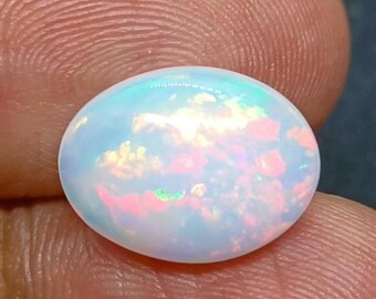Könnte beinhalten: Ein ovaler Opal-Cabochon-Edelstein mit einer durchscheinenden weißen Basis und lebhaften Blitzen in Pink, Orange und Grün, die sich über den gesamten Stein erstrecken. Der Edelstein hat eine glatte, polierte Oberfläche und ein gewölbtes Profil.