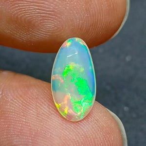 Peut inclure: Une pierre précieuse opale ovale vibrante. L'opale présente un jeu de couleurs avec des éclairs de vert, de bleu, de jaune et d'orange dans une base blanche laiteuse translucide. La pierre est tenue entre deux doigts.