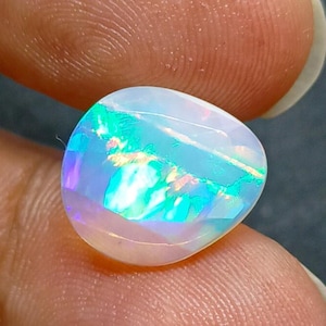 May include: A polished, oval-shaped opal gemstone. The opal displays a play of color with iridescent flashes of blue, green, and purple. The stone has a translucent, milky white base color. The gemstone is held between two fingers.