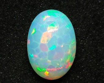 May include: An oval opal gemstone with a unique pattern. The opal has a milky white base with flashes of green, orange, and red. The gemstone is set against a dark, textured background.