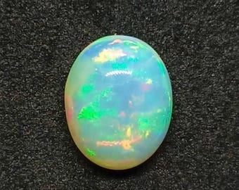Könnte beinhalten: Ovale Opal-Edelstein mit milchig-weißer Basis und lebendigen grünen, blauen und rosa Blitzen. Der Edelstein ist vor einem dunklen, strukturierten Hintergrund platziert, der seine irisierenden Eigenschaften hervorhebt. Ein natürlicher Edelstein.