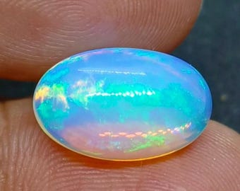 Könnte beinhalten: Ein ovaler Opal-Edelstein, der ein faszinierendes Farbspiel zeigt. Die Oberfläche des Steins reflektiert ein Spektrum an Farbtönen, darunter Blau, Grün und Gelb, wodurch ein faszinierender visueller Effekt entsteht. Der Opal wird zwischen Fingern gehalten, was seine natürliche Schönheit hervorhebt.