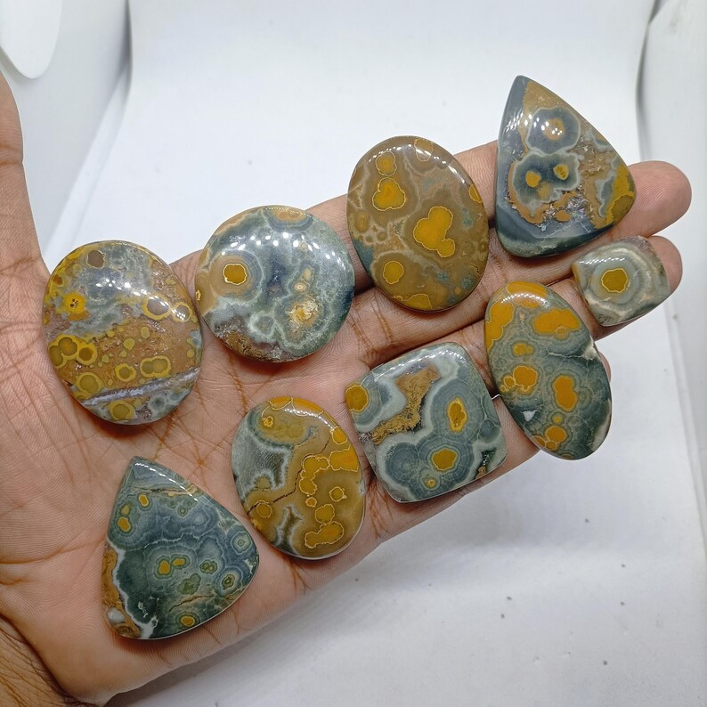 May include: A collection of nine polished ocean jasper cabochons in various shapes, including oval, round, square, and teardrop. Each stone features a distinct pattern of circular yellow and orange orbs set against a background of muted grey, green, and brown tones.