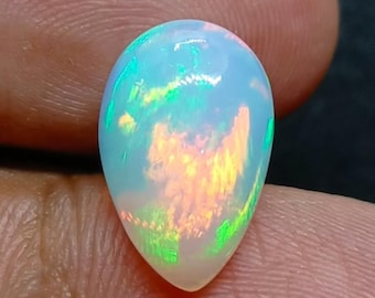 May include: A pear-shaped opal gemstone with a milky white base. The opal exhibits a play-of-colour effect with flashes of green, yellow, and orange. The gemstone is held between fingers. The opal is a beautiful example of its type.