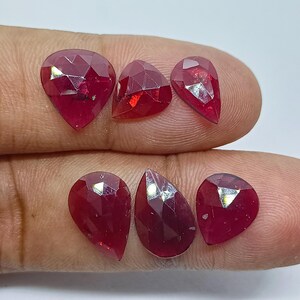 May include: Six faceted, teardrop-shaped rubies in varying sizes. The gemstones are a deep, rich red color, with multiple facets catching the light. The rubies are displayed against a neutral background.