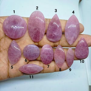 Può includere: Undici cabochon di quarzo rosa lucidati in varie forme, tra cui rotonda, ovale, a goccia e a navetta, ciascuno numerato con una cifra nera da 1 a 11. Le pietre presentano una tonalità rosa chiaro traslucida con naturali texture cristalline.