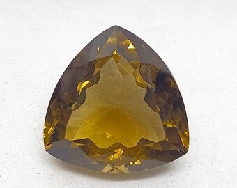 Op de afbeelding: Een gefacetteerde, driehoekig geslepen edelsteen met een diepe goudbruine, doorschijnende kleur. De steen heeft een symmetrisch, veelzijdig oppervlak dat licht reflecteert, wat zorgt voor een helder, gepolijst uiterlijk met scherpe randen.