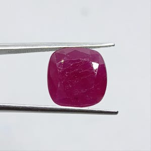 Puede incluir: Una piedra preciosa de rubí con corte cojín, de tono rojo magenta intenso y con inclusiones internas visibles. La piedra facetada está sujeta por unas pinzas de metal plateado, mostrando su superficie pulida y su forma geométrica.