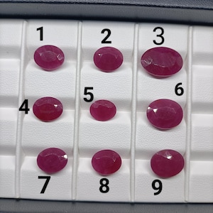 Peut inclure: Une collection de neuf rubis rouge rosé profond, taillés en ovale et disposés dans un plateau de présentation blanc. Chaque pierre précieuse est numérotée de un à neuf, avec des surfaces facettées reflétant la lumière sur leurs corps polis.