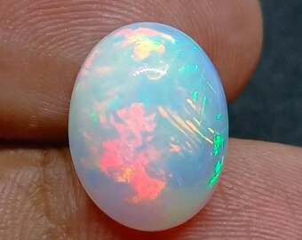 Könnte beinhalten: Ovale Opal-Edelstein mit einer schillernden, irisierenden Oberfläche. Der Stein zeigt Blitze in Rot, Orange, Grün und Blau. Der Opal wird zwischen zwei Fingern gehalten, was seine polierte Textur und das lebendige Farbspiel hervorhebt.