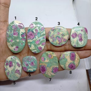 Könnte beinhalten: Acht polierte Cabochon-Edelsteine mit einem gesprenkelten Muster aus leuchtend magentafarbenen Einschlüssen, eingebettet in eine Matrix aus hellgrünem, cremefarbenem und blaugrünem Mineralmaterial. Jeder Stein ist zur Identifizierung nummeriert.