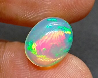 Könnte beinhalten: Ein polierter, ovaler Opal-Edelstein. Der Opal zeigt eine transluzente, milchig-weiße Grundfarbe mit lebendigem internem Farbspiel, einschließlich rosa, grüner und gelber Blitze. Der Edelstein wird zwischen zwei Fingern gehalten.