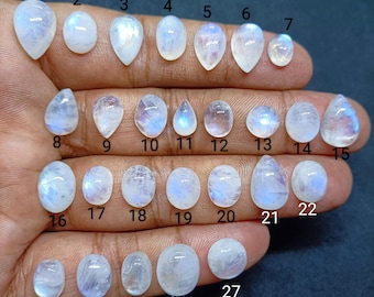 May include: A collection of twenty-seven polished moonstone cabochons in various shapes including oval, pear, and round. Each gemstone displays a translucent white appearance with a distinct blue adularescence. Each stone is numbered from 1 to 27.