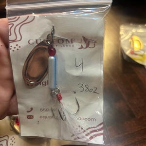 May include: A fishing lure in a clear plastic bag. The lure has a copper-colored blade, a red bead, and a white and blue body. The bag is labeled "Custom Lures" and has the number "4" and "38oz" written on it.
