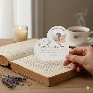 May include: A heart-shaped memorial card with the name "Pauline Archer" and a photo, held over an open book. The card features doves and floral accents, with text in French. A lit candle and a cup of tea are in the background.